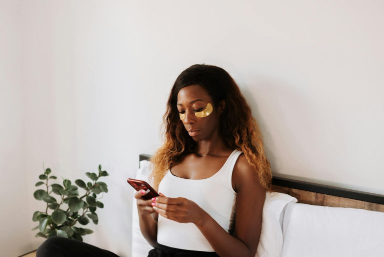 A woman relaxes in bed applying under eye masks while using her smartphone.